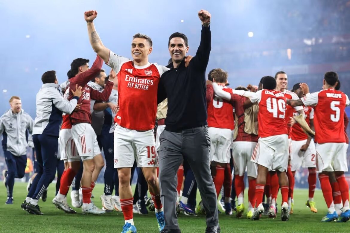 Arsenal players and coach celebrate a victory on the pitch with arms raised in triumph as teammates hug and cheer in the background.