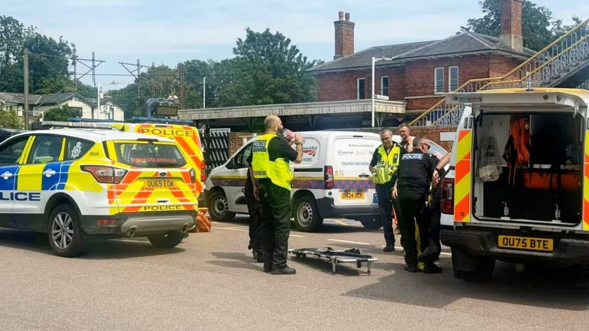 Multiple emergency vehicles (police cars and ambulance) at an incident scene, with officers in high-visibility vests standing around a wheeled stretcher on the pavement.