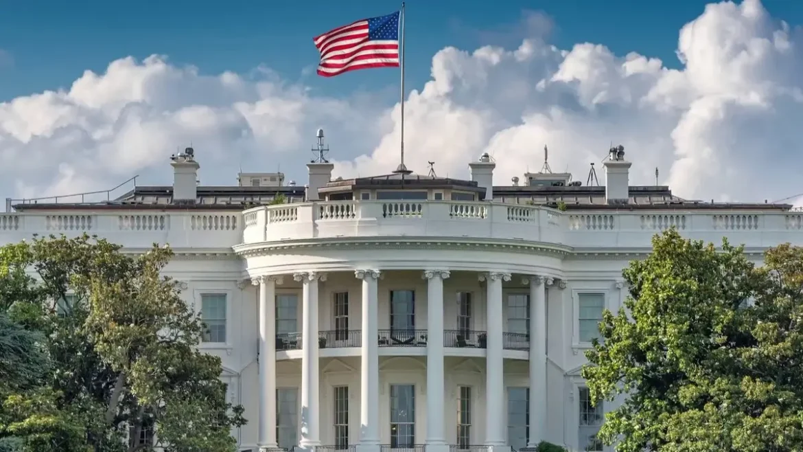 Front view of the White House with a flag flying atop and a blue sky with clouds behind it, surrounded by trees.