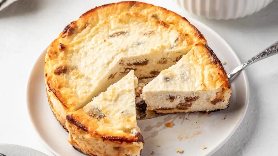 Round cheesecake with a golden crust and visible raisins on a white plate; a slice has been cut and served nearby.