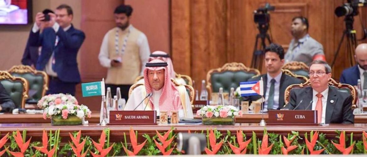 Saudi official and Cuban delegate sit at a formal conference table with flags, flowers, and cameras in the background.