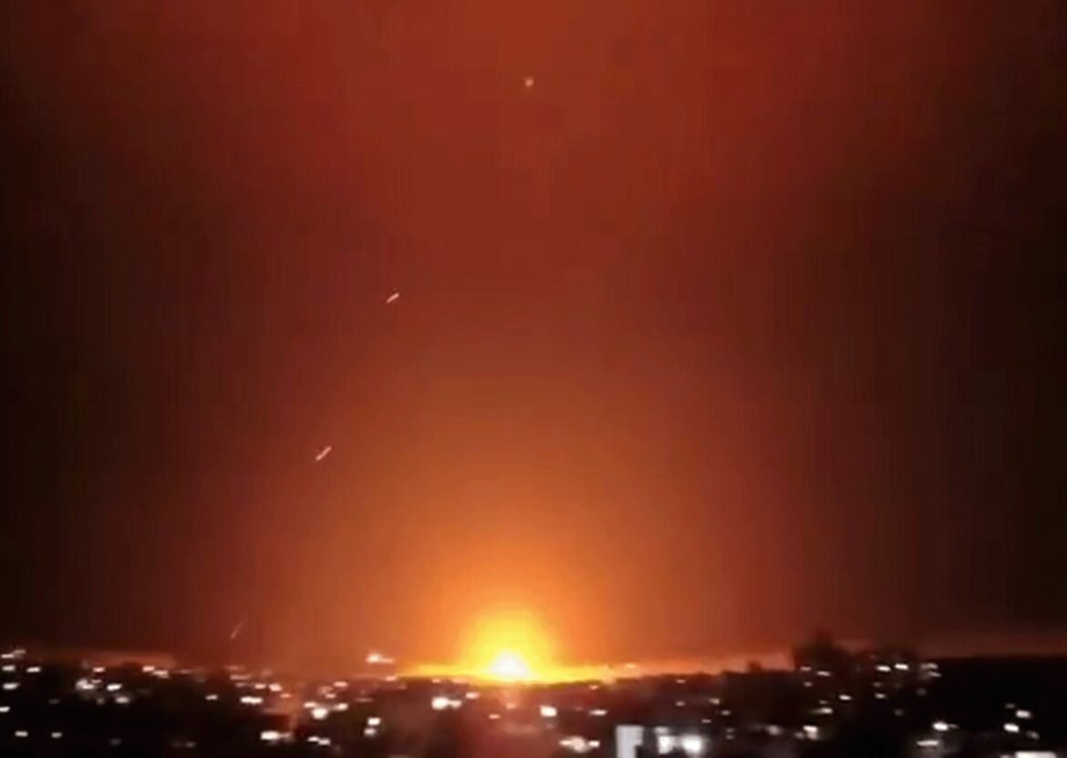 Nighttime cityscape lit by a large orange explosion on the horizon.