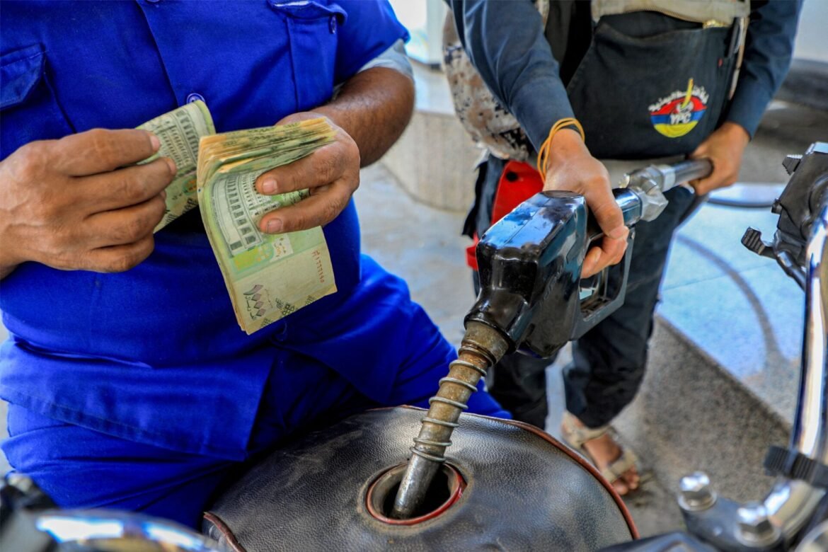 Man in blue uniform counts a stack of cash at a gas station, with a fuel nozzle near a motorcycle being held by another person.