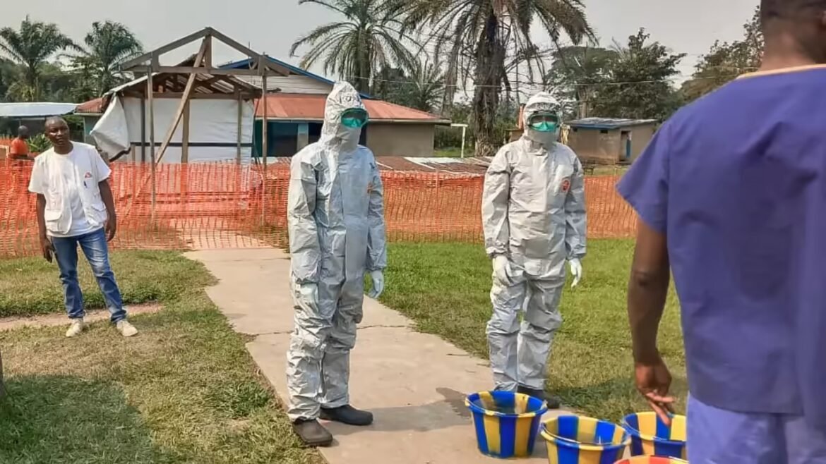 Two people in full hazmat suits and green goggles stand on a path near a fenced area with orange mesh, buckets at their feet.
