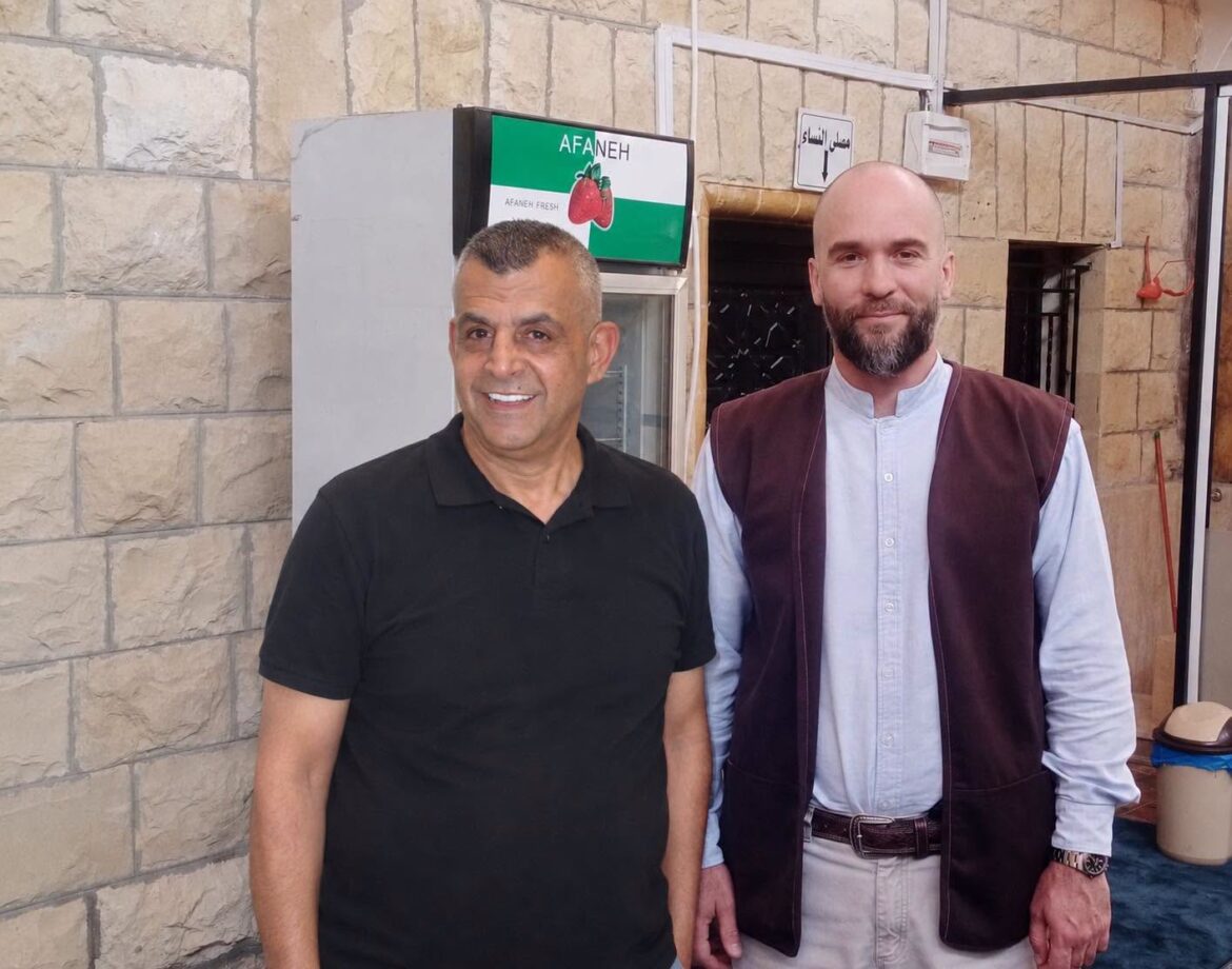Two men smile for the camera indoors, standing beside a refrigerator against a stone wall, one in a black polo shirt and the other in a brown vest over a light shirt.