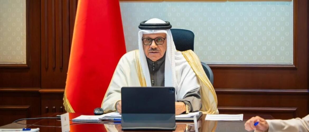 Senior man in traditional white robe and head covering sits at a desk with a laptop, flag in the background, looking at the camera.