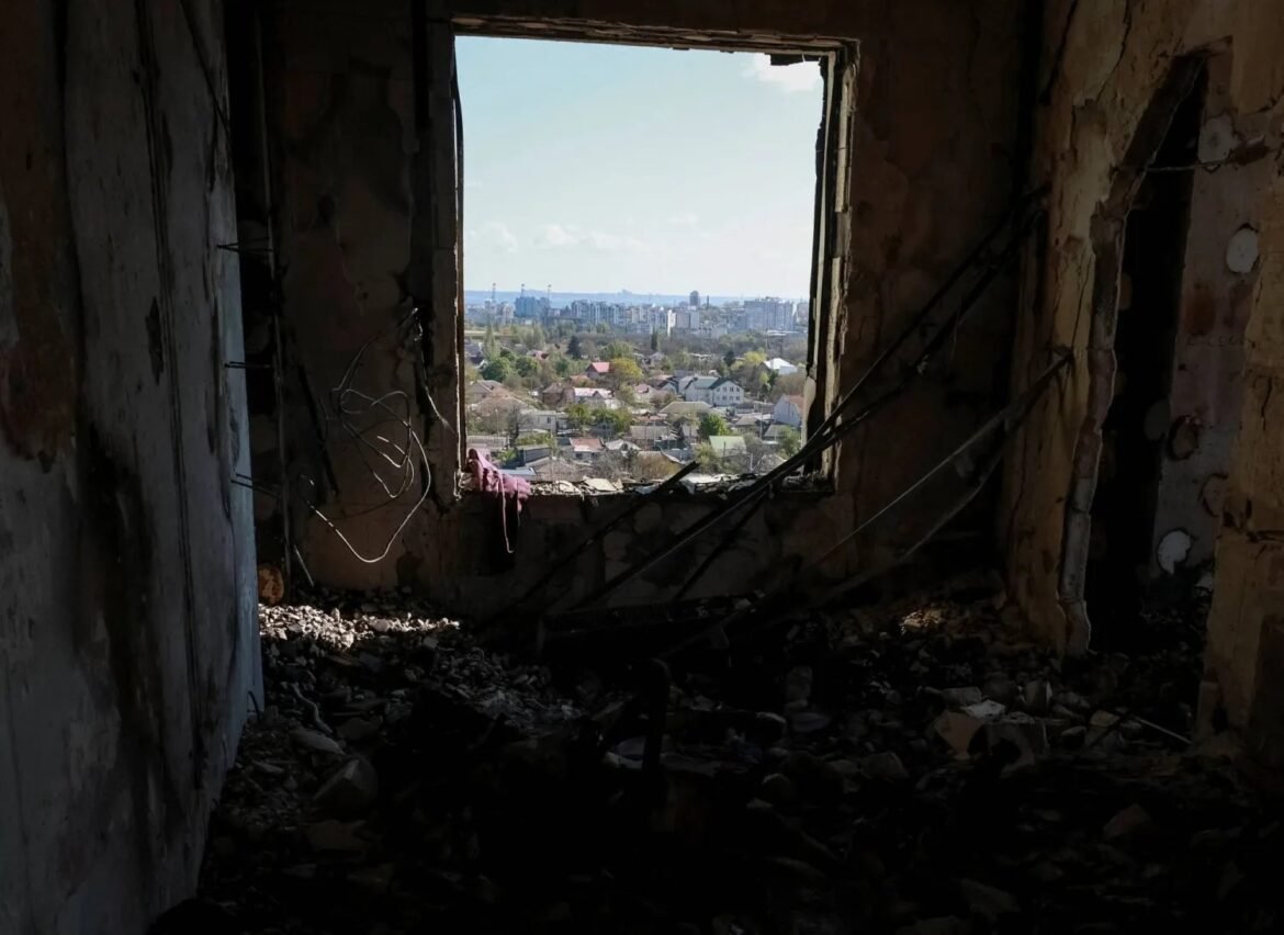Ruined interior of a building with a large window opening showing a residential cityscape outside, on a sunny day.