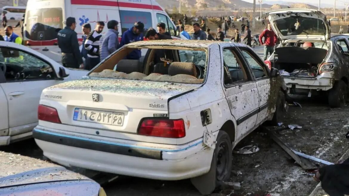 White sedan severely damaged in a multi-vehicle crash, debris on the ground, with an ambulance and bystanders in the background.