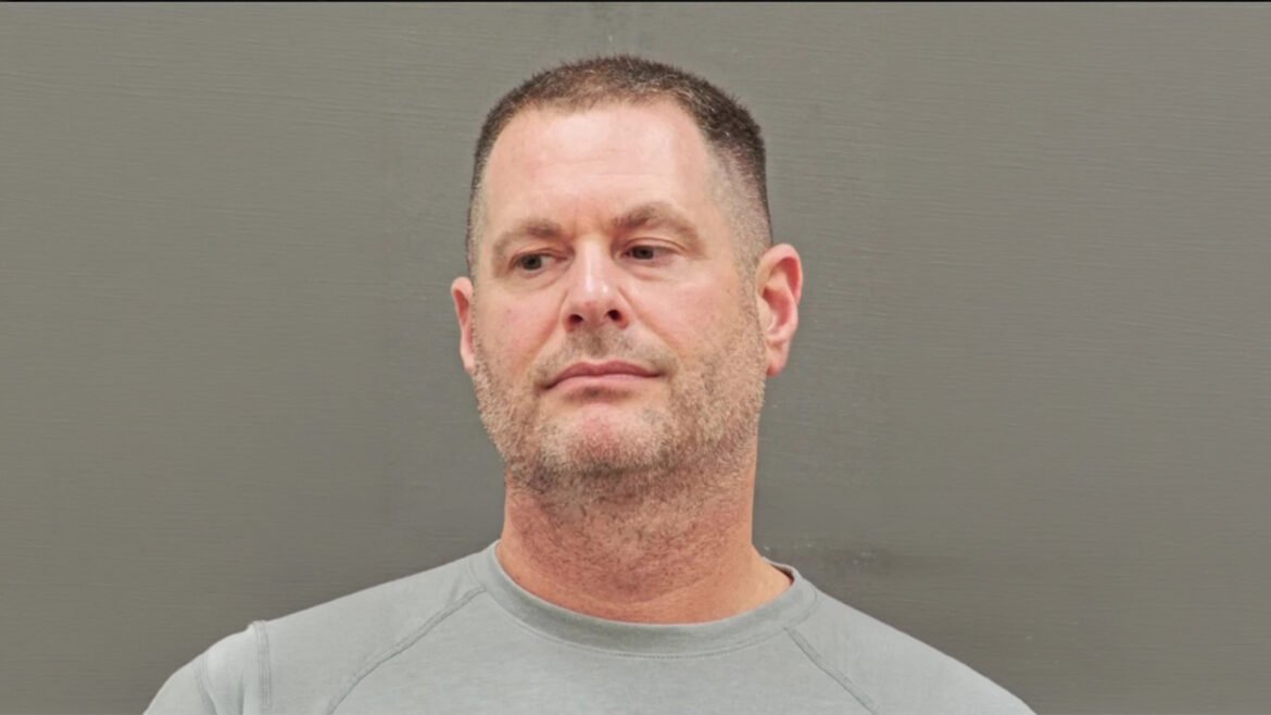Mugshot-style photo of a middle-aged man with short dark hair and stubble, wearing a light gray shirt, against a gray wall.
