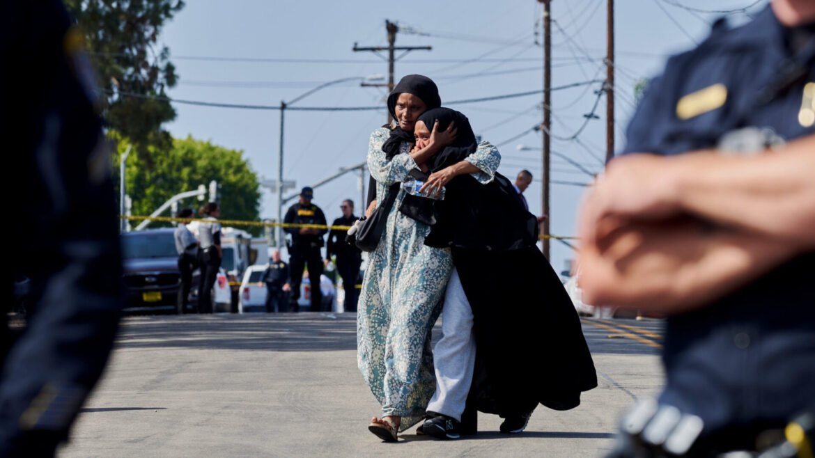 Two women wearing hijabs hug in the street while police and yellow tape frame the background.