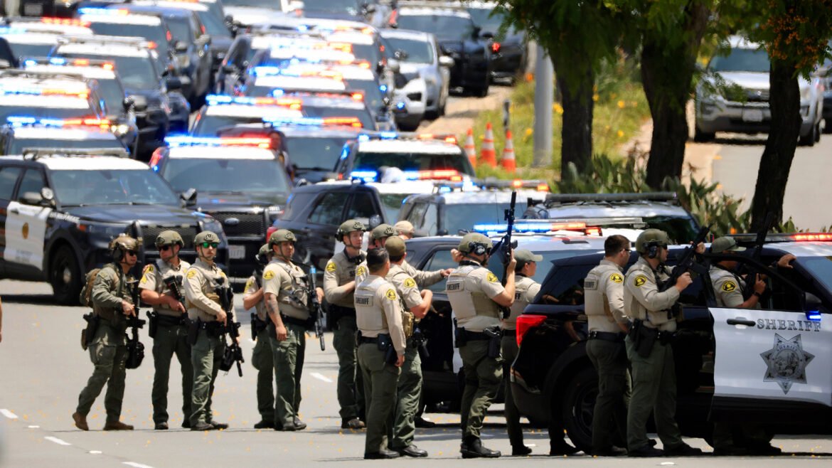 Line of sheriff's deputies in tactical gear with rifles standing on a street beside patrol SUVs with flashing lights.