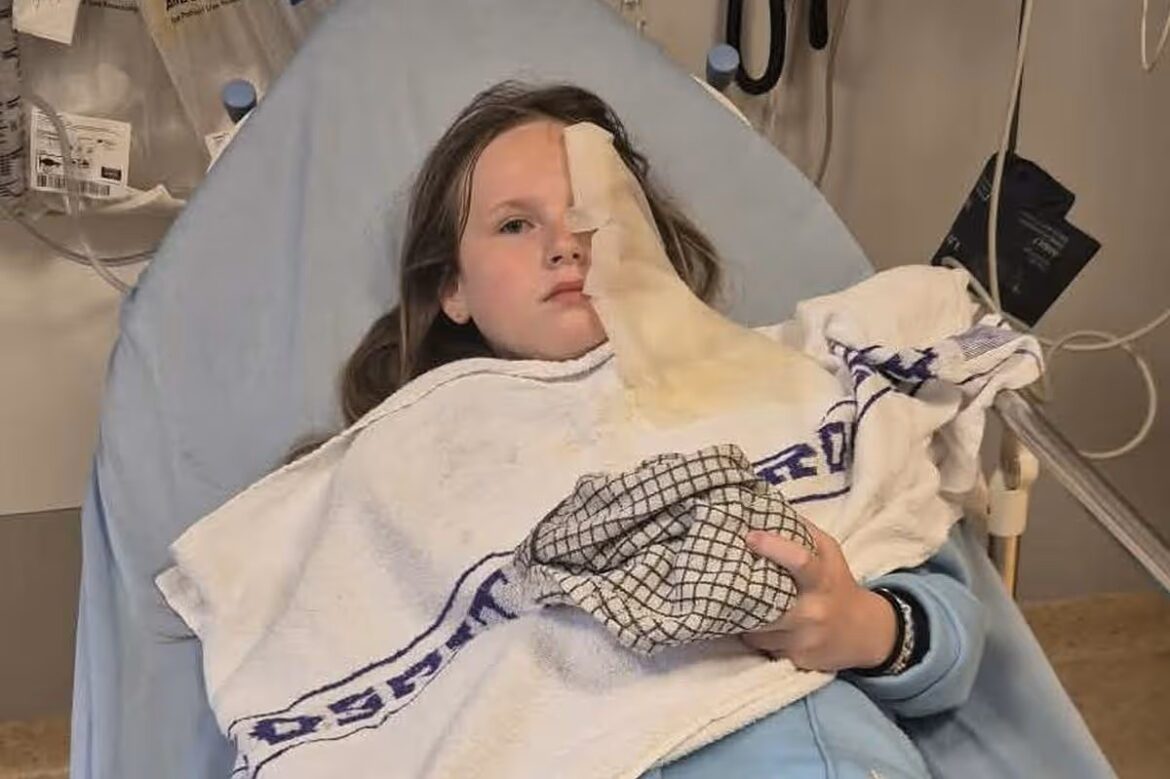 Girl in a hospital bed with a large facial bandage covering her left eye, resting under a towel and blanket in a medical room.