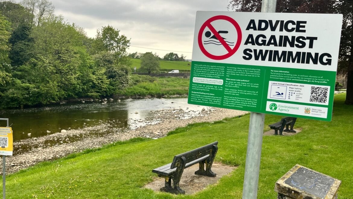 Large sign reading 'Advice Against Swimming' by a river in a park with benches nearby.