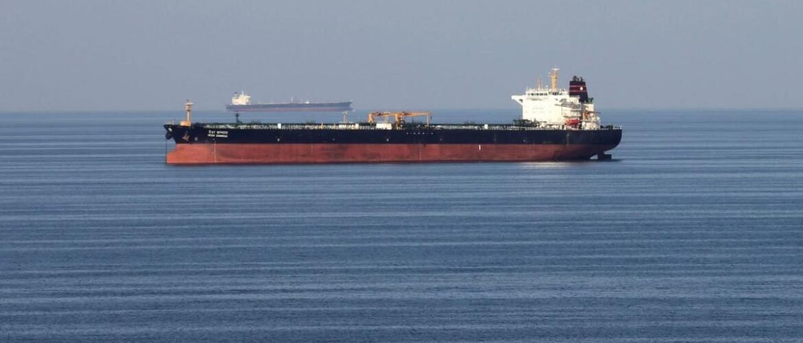 Large bulk carrier with a red hull sails on calm blue sea; a smaller vessel is visible in the distance.