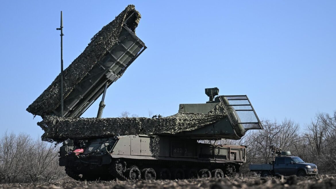 Self-propelled missile launcher with camouflage netting, elevated launcher rail extended over barren field under clear blue sky (military vehicle)