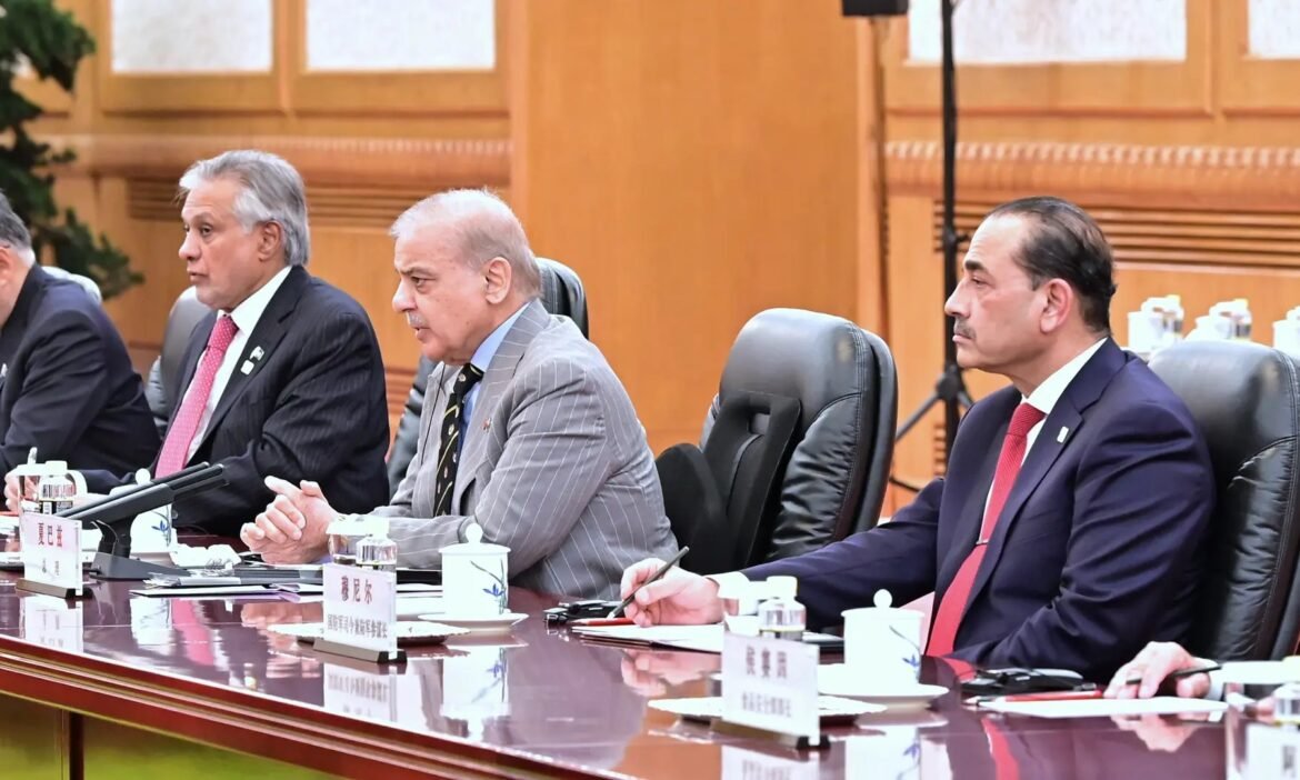 Three men in suits sit at a polished conference table during a formal meeting, with microphones and nameplates visible on the table.
