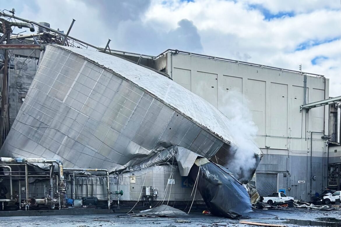 Industrial facility with a large tilted metal silo/duct collapsed onto debris and a blue tarp, steam rising from the wreckage