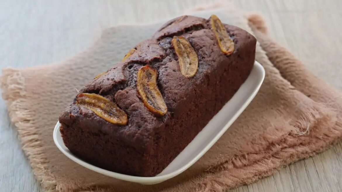 Chocolate banana bread loaf on a white dish, topped with banana slices and resting on a beige cloth.