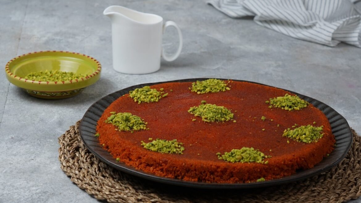 Round red cake topped with small pistachio mounds on a dark plate, with a white jug and a green pistachio bowl in the background.