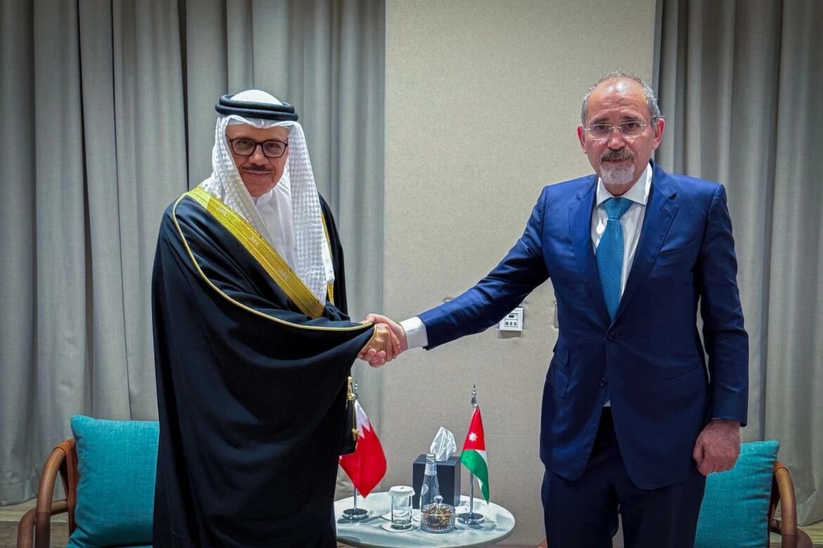 Two men shake hands in a formal meeting: one in a traditional Arab robe and headscarf, the other in a blue suit, with small tables and flags nearby.