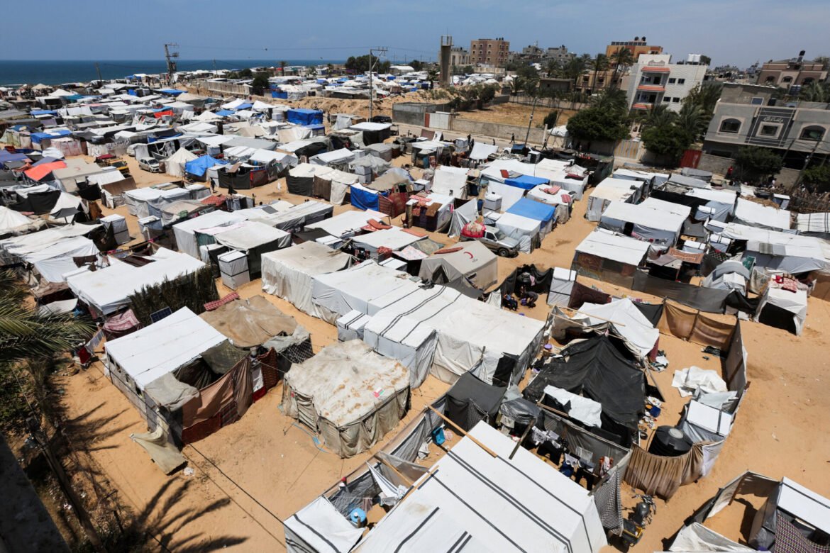 Overhead view of a crowded sea-side refugee camp with rows of white tents and makeshift shelters on sandy ground.