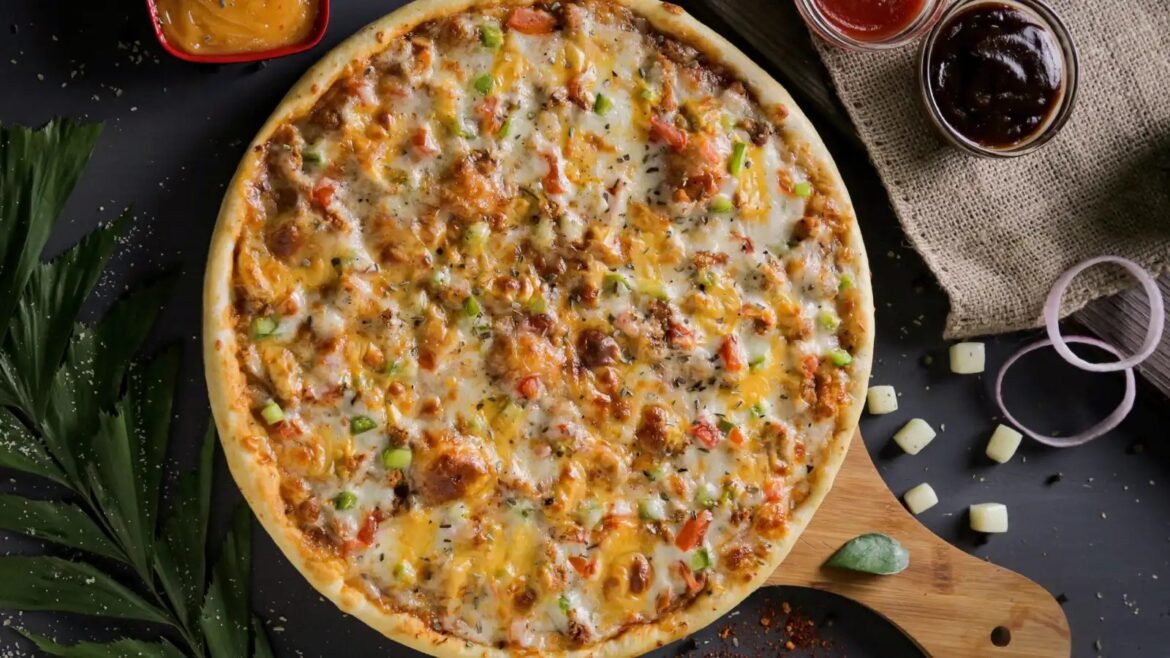 Cheese pizza topped with peppers and onions on a wooden pizza board, with sauce jars nearby on a dark surface.