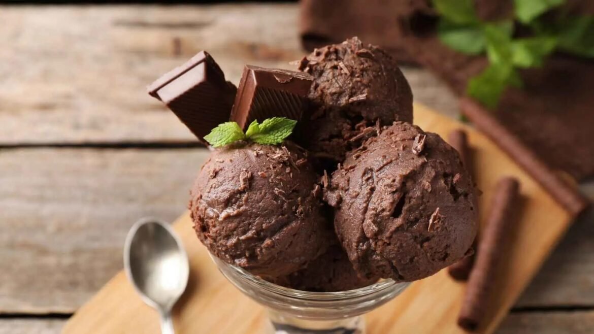 Three scoops of chocolate ice cream with chocolate chunks and a mint leaf, served in a glass bowl on a wooden table.