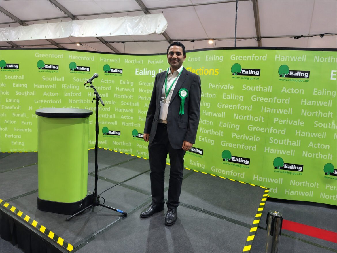 Man in a dark suit with a green rosette stands on a small stage beside a green podium and microphone.