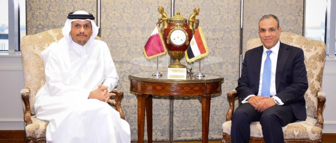 Two men sit in ornate chairs on either side of a round table with flags and a clock-highlighted centerpiece, indicating a formal meeting between Qatar and Egypt representatives.