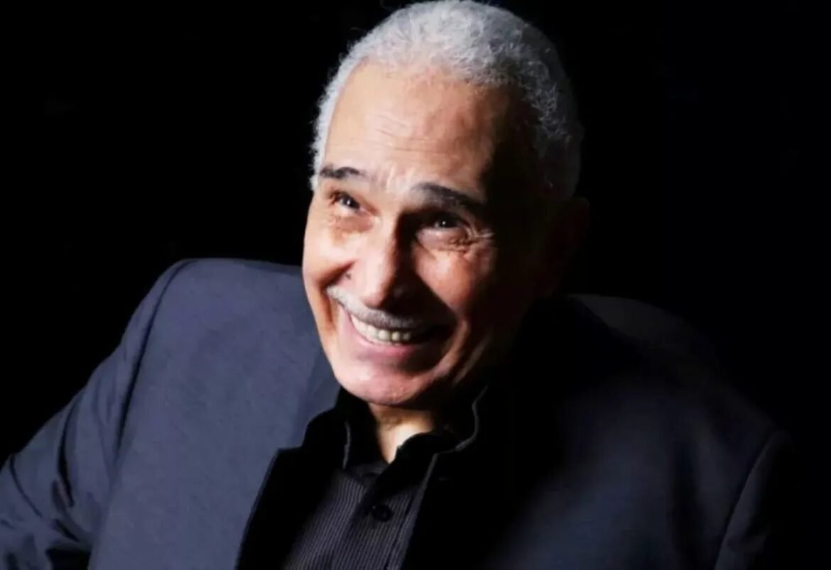 Smiling older man with gray hair wearing a dark blazer against a black background.