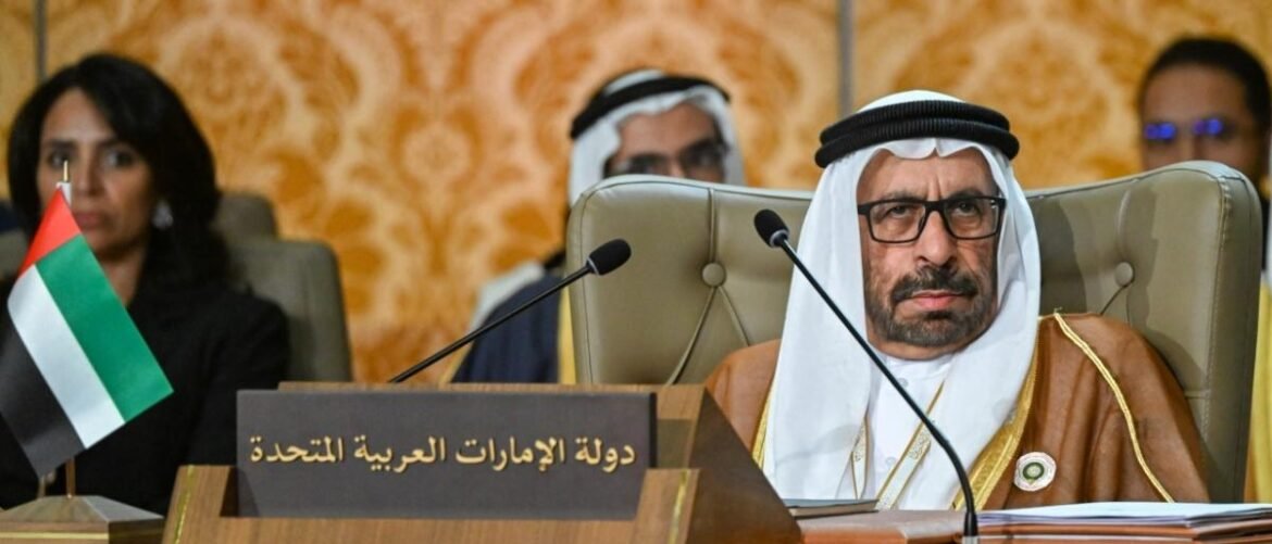 رجل خليجي مرتدياً ثوباً وغطاء رأس يجلس في مؤتمر أمام لوحة مكتوبة بالعربية: 'دولة الإمارات العربية المتحدة' وعلم الإمارات ظاهر على الطاولة рядом.