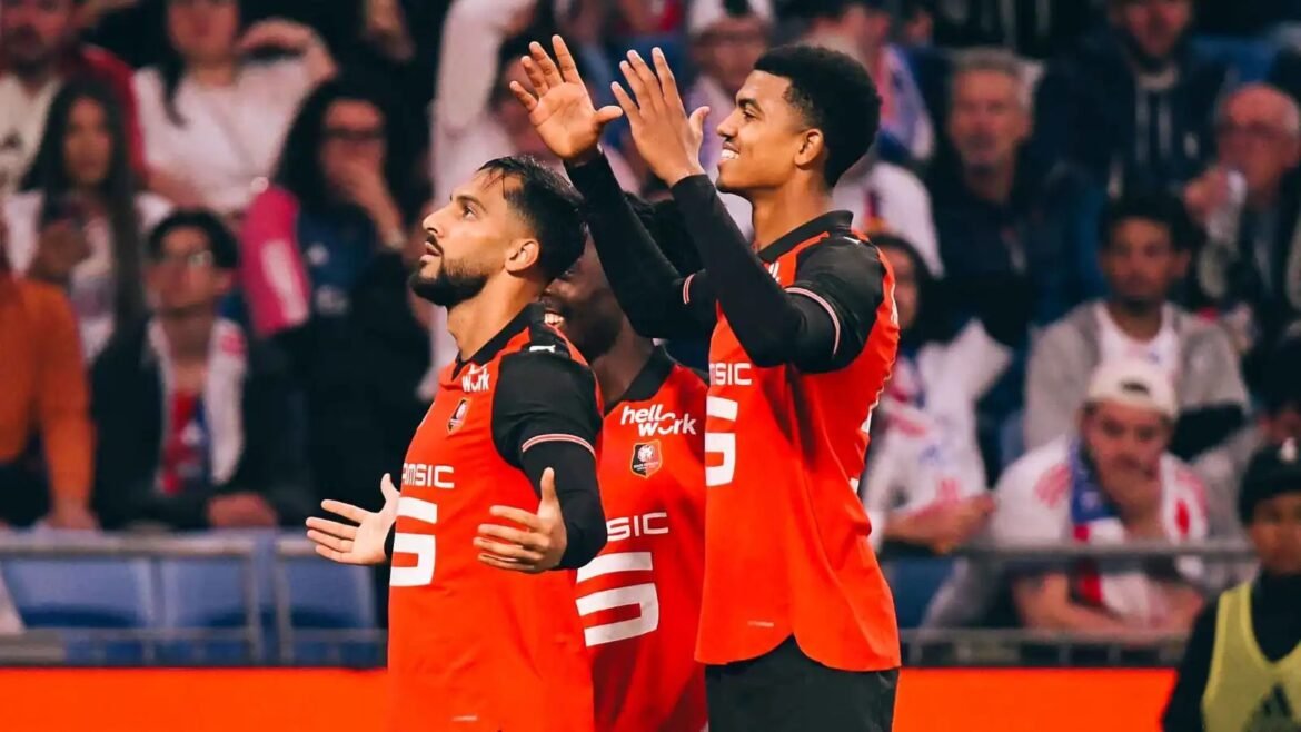 Two soccer players in red and black uniforms celebrate on the field with raised hands as the crowd cheers behind them.