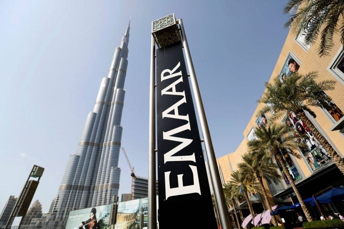 Tall Burj Khalifa skyscraper visible behind a vertical EMAAR sign at a sunny outdoor shopping area with palm trees and storefronts on the right.