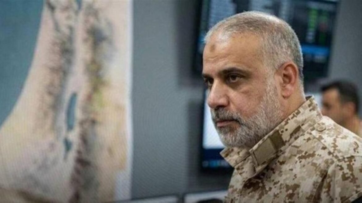 Side profile of a bearded man in desert camouflage military attire in a control room with monitors in the background.