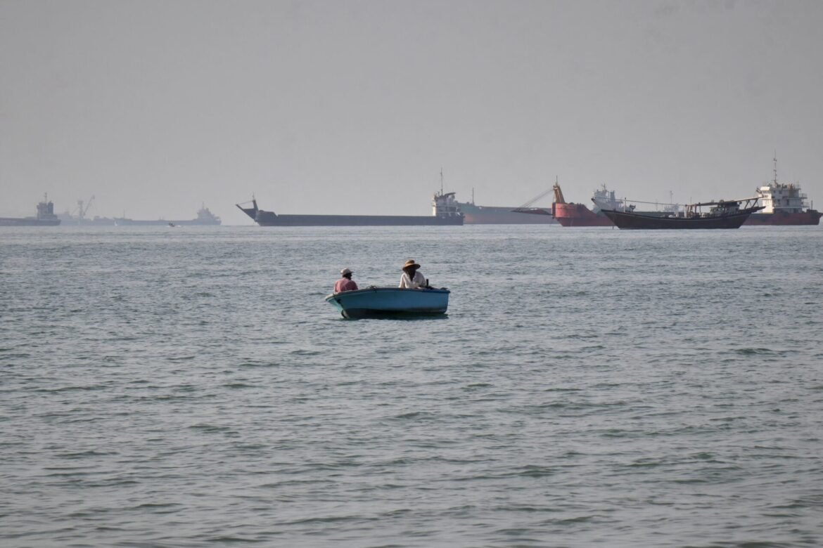 Two people in a small blue boat on calm water, with large cargo ships and cranes visible in the hazy harbor in the distance.