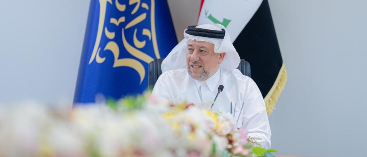 A man in a white thobe and headscarf speaks at a conference with flags in the background and flowers in the foreground.