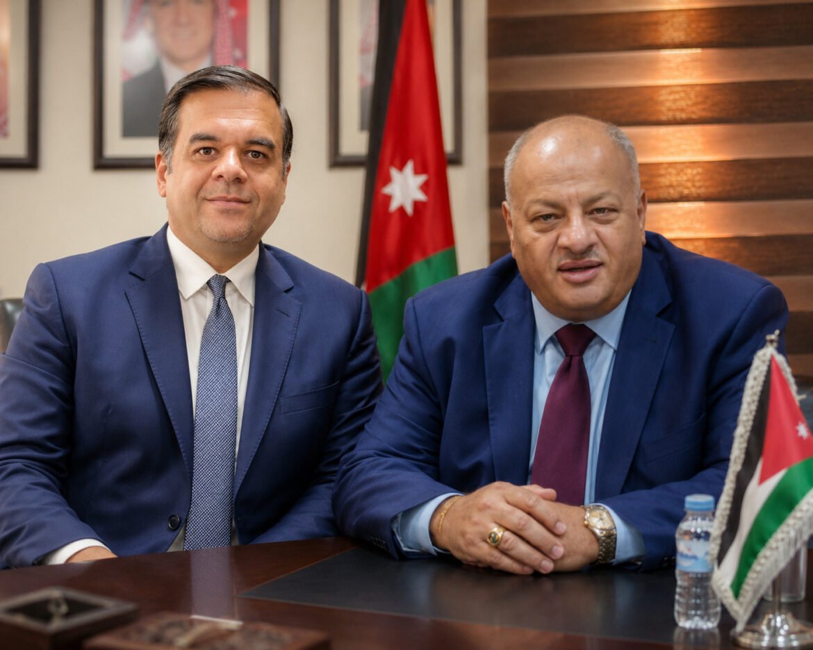 Two men in navy suits sit at a conference table in a formal office, with a Jordan flag in the background.
