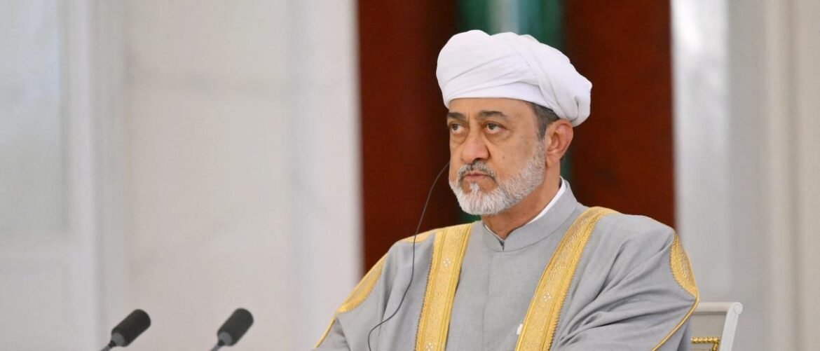 Man in a white turban and grey ceremonial robe with gold trim seated at a table with two microphones in front.