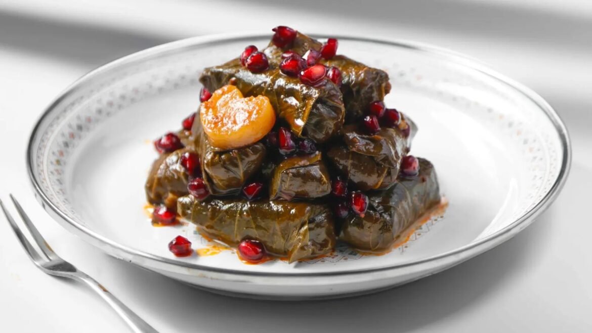Stack of grape leaf dolmas drizzled with sauce and sprinkled with pomegranate seeds on a white patterned plate.