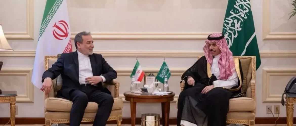 Two men in formal attire sit on ornate chairs in a luxurious room, with Iran and Saudi flags on a small table between them, suggesting a diplomatic meeting.