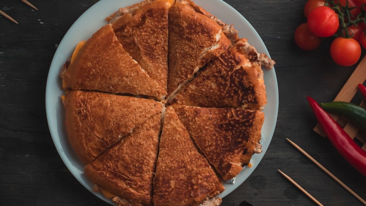Sesame-crusted flatbread sandwich cut into wedges on a light blue plate, with tomatoes nearby on the dark surface.