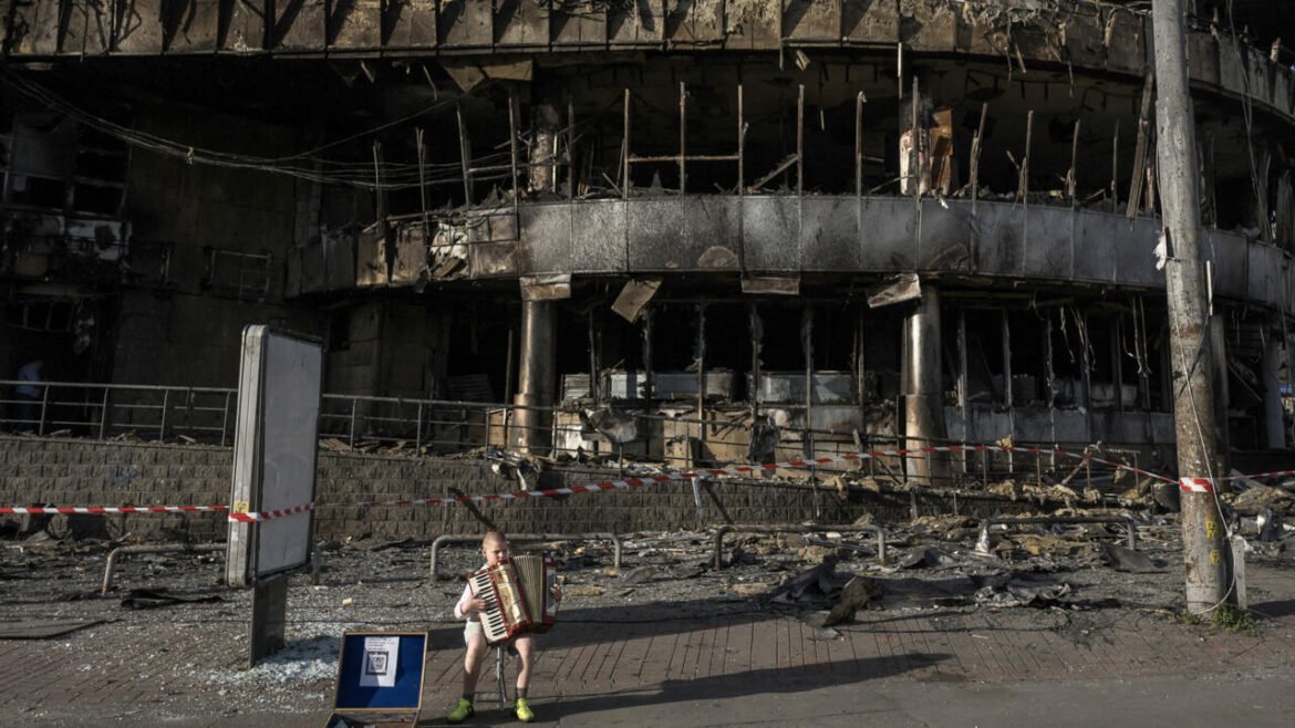 Boy sits on a small chair playing an accordion in front of a severely burned, rubble-strewn building with caution tape nearby start of street scene