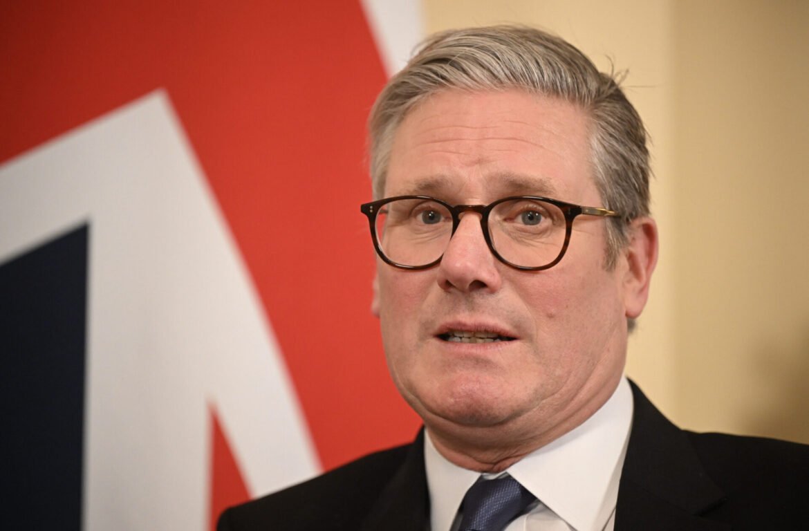 Close-up of a middle-aged man wearing glasses and a suit, speaking at an event with a red-and-white backdrop behind him.