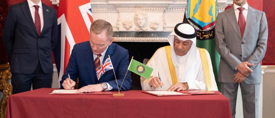 Two men sign documents at a formal signing ceremony, with flags visible behind them and two observers standing nearby.
