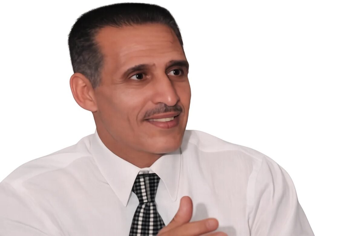 Man in a white dress shirt and checkered tie smiling and gesturing with his hand, looking to the right against a white background.