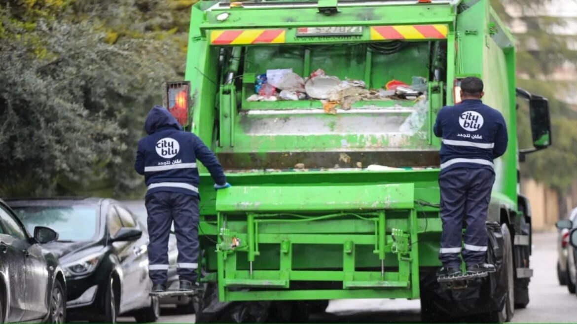 Two city sanitation workers in reflective uniforms stand on the rear platform of a bright green garbage truck, sorting and collecting trash on a street at curbside edge.