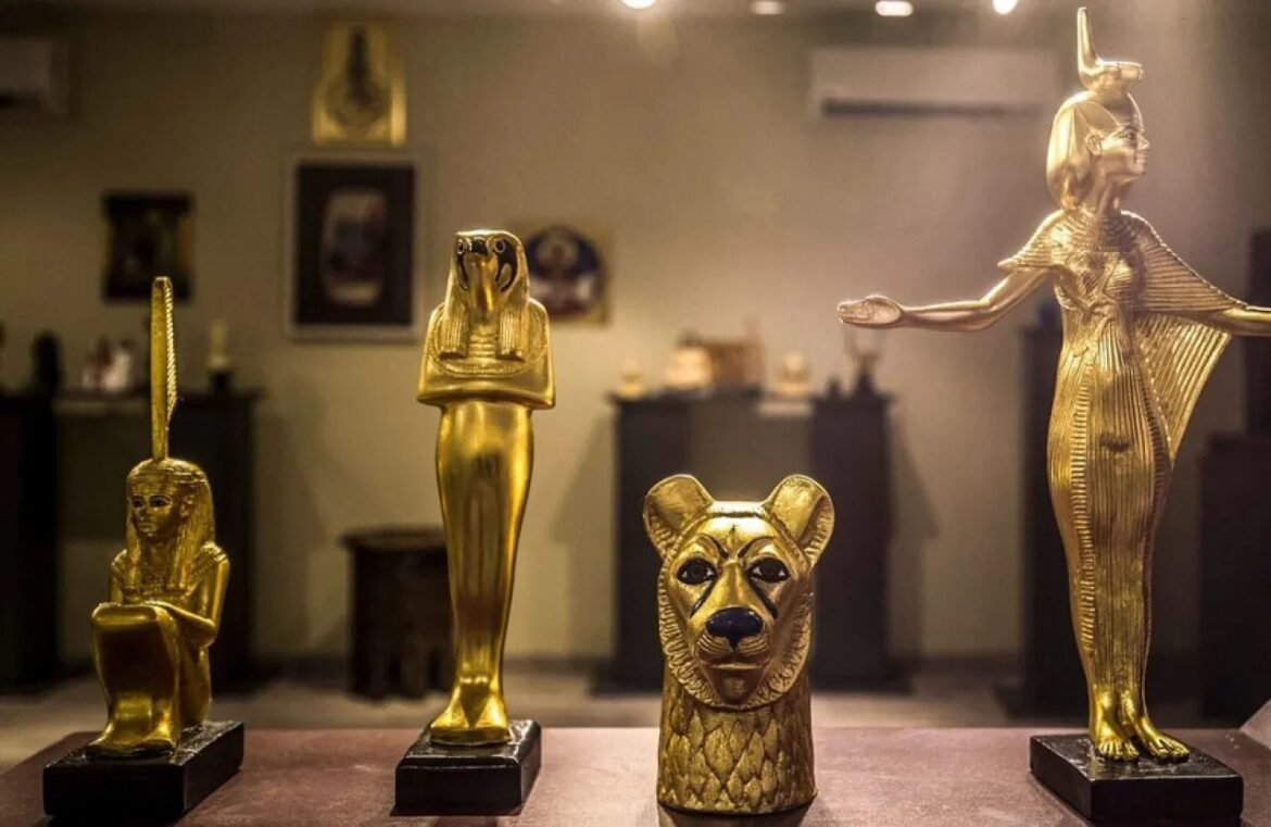 Gold-toned display of ancient-style statues: a seated figure, a tall standing figure, a lion-headed sculpture, and a winged deity on pedestals in a museum case.