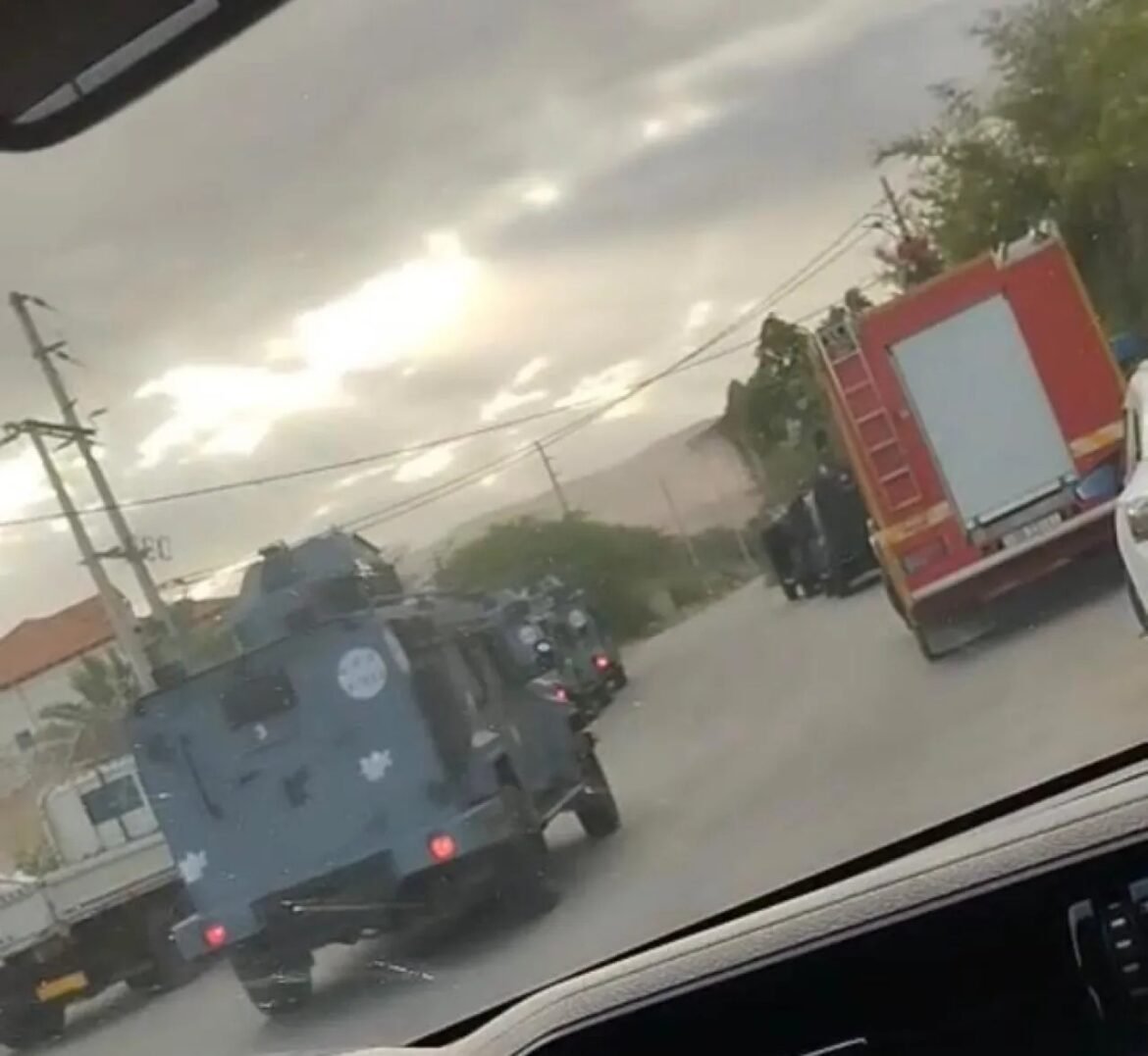 Rear view of a blue armored vehicle on a wet street with a red fire truck to its right during a rainstorm, seen from a car window.