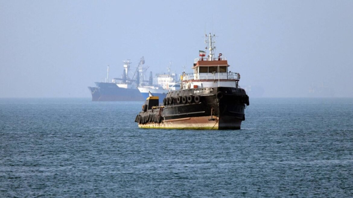 Tugboat sailing on calm blue water with larger ships blurred in the hazy distance behind it.