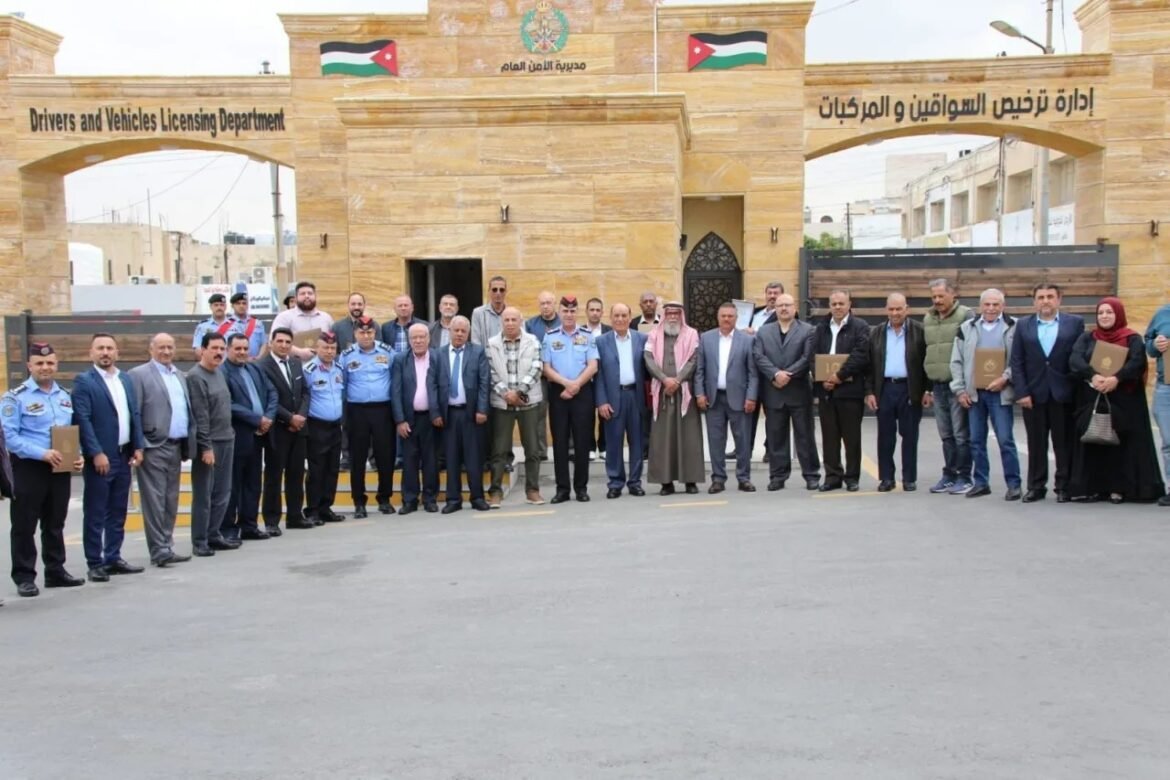 Group of officials and staff posing outside the Drivers and Vehicles Licensing Department building, with Jordanian flags overhead.